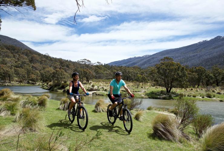 Two people cycling along a grass track beside a river surrounded by greenery at The Diggings, Kosciuszko National Park, New South Wales © Destination NSW