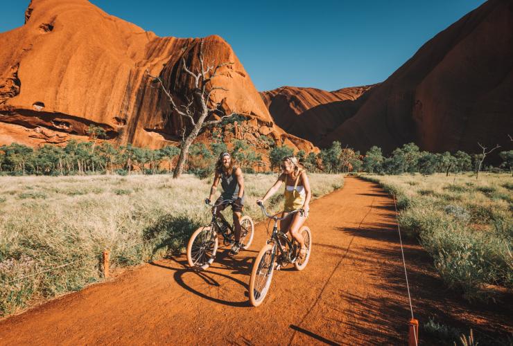 Two people laughing while cycling along a red sand track with the towering formation of Uluru behind them in Uluru-Kata Tjuta National Park, Northern Territory © Tourism NT/Laura Bell