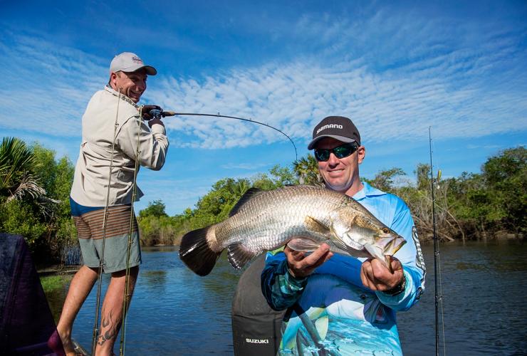A man holding a fish as another man casts a rod into a waterway, Dhipirri Barra & Sportfishing, Arnhem Land, Northern Territory © Shaana McNaught