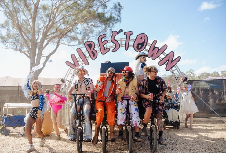 A group of friends dressed in colourful costumes for a festival while posing under a sign reading "vibestown", Party in the Paddock, Carrick, Tasmania © Cameron Jones