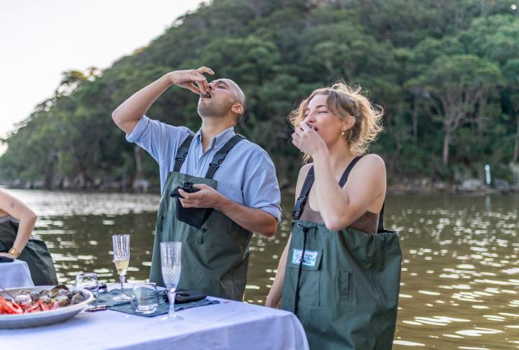 Two people standing in a river at a table eating freshly shucked oysters, Sydney Oyster Farm Tours, Hawkesbury, New South Wales © Tourism Australia