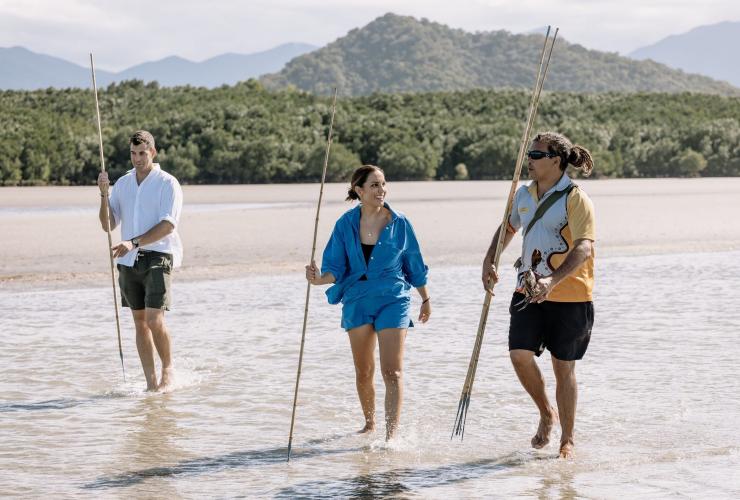 Two guests walking across a mudflat with a local Aboriginal guide while holding spears and a mudcrab, Walkabout Cultural Adventures, Mossman, Tropical North Queensland © Tourism Australia