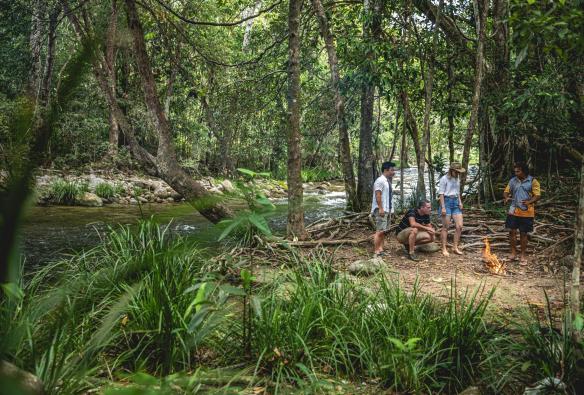 A group of people standing around a campfire in a rainforest beside a river with Walkabout Cultural Adventures, Port Douglas, Tropical North Queensland, Queensland © Tourism and Events Queensland