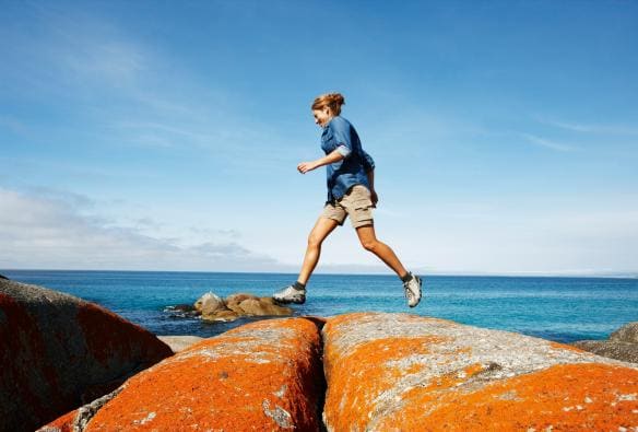 A person hopping along orange-lichen covered rocks by the ocean, Binalong Bay, Bay of Fires Conservation Area, Tasmania © Tourism Australia