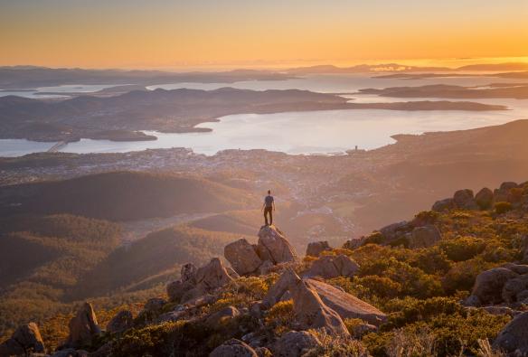 A person standing on a mountain peak overlooking a harbour city during sunrise, kunanyi/Mount Wellington Summit, Hobart, Tasmania © Justin Hyde