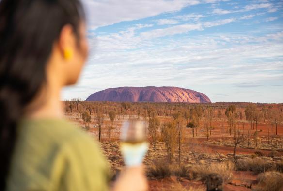 A person holding a glass on wine and looking towards a large red rock monolith, Sounds of Silence, Uluṟu, Northern Territory © Tourism NT/Helen Orr