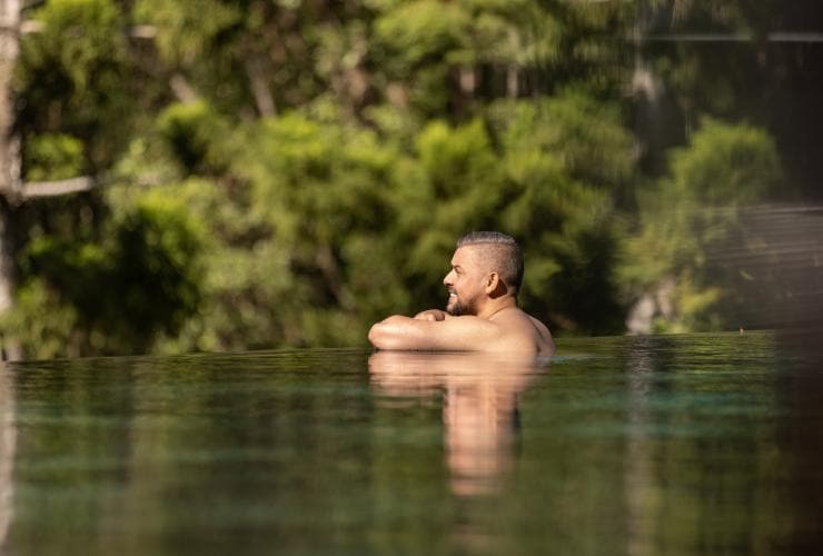 A male guest leaning on the edge of the infinity pool surrounded by trees at Gwinganna Lifestyle Retreat, Gold Coast, Queensland © Tourism Australia