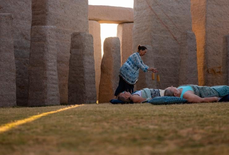 Guests laying down on yoga mats and pillows on grass amid stone structures as an instructor waves instruments above them during a retreat with Untamed Escapes, Esperance, Western Australia © Tourism Australia