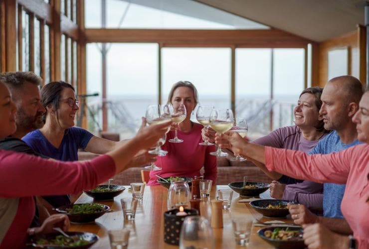 A group of people drinking glasses of white wine over a meal in a lodge, Bay of Fires Signature Walk, Bay of Fires Conservation Area, Tasmania © Tourism Australia