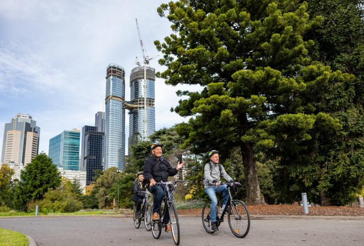 Three people riding bikes along a quiet path through a leafy park with the city skyline behind them during a tour with Melbourne by Bike, Melbourne, Victoria © Tourism Australia