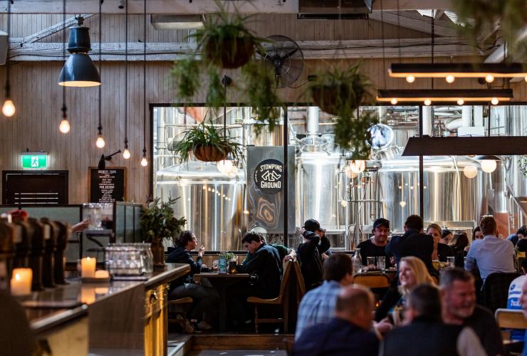 A crowded venue filled with people smiling and drinking beer at tables beside large windows leading to views of brewing equipment at Stomping Ground Brewery Co., Collingwood, Victoria © Stomping Ground Brewery Co.