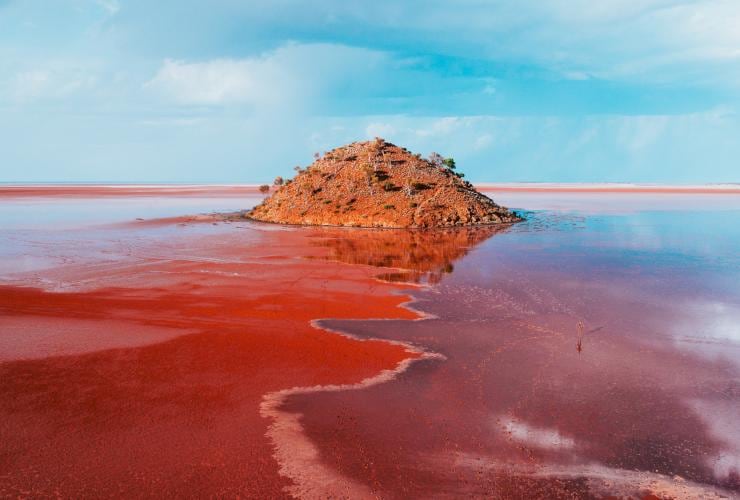Aerial view over a vibrant pink lake with a tall rocky formation in the middle and a sculpture to the side at Lake Ballard, Menzies, Golden Outback, Western Australia © Australia’s Golden Outback
