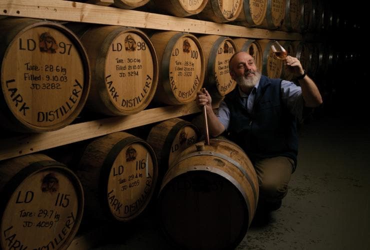 Man kneeling beside a wall of oak barrels admiring a freshly poured glass of liquor at Lark Distillery, Hobart, Tasmania © Osborne Images