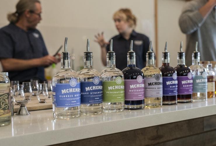 A range of colourful spirits lined up along a table with guests chatting in the background at McHenry Distillery, Tasman Peninsula, Tasmania © Tourism Australia