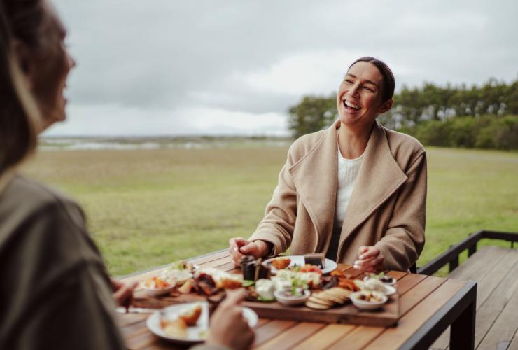 Two people smiling over a meal of native ingredients enjoyed while overlooking sweeping green fields, Tae Rak Aquaculture Centre Café, Budj Bim National Park, Great Ocean Road, Victoria © Tourism Australia