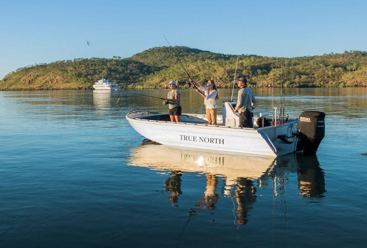 Three people fishing from a small boat on a glassy waterway with a larger cruise ship in the background, True North, The Kimberley, Western Australia © True North