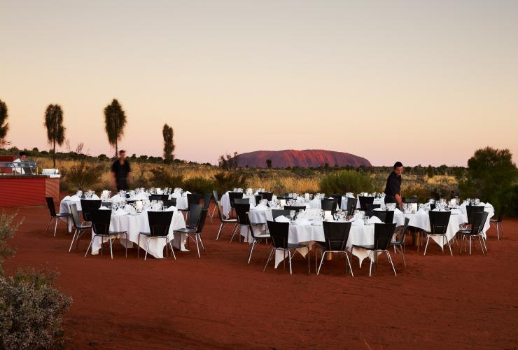 Tables and chairs set in an outback, red dirt landscape with a large rock monolith in the distance, Sounds of Silence, Uluṟu-Kata Tjuṯa National Park, Red Centre, Northern Territory © Tourism Northern Territory