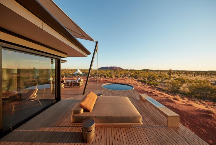 Exterior of accommodation at Longitude 131 with two people seated on a private deck with lounges and plunge pool looking at an unobstructed view of Uluru in the background in Yulara, Northern Territory © Baillie Lodges