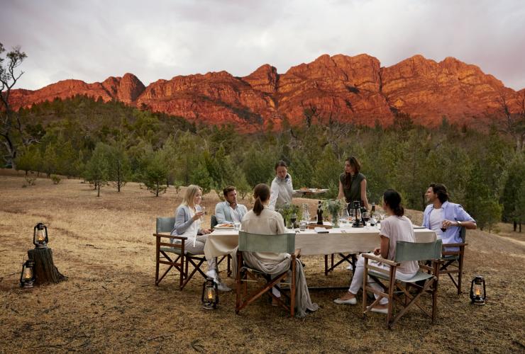 A group dining outdoors on a grassy plain surrounded by trees with mountain peaks glowing red in the distance at Arkaba Conservancy, Flinders Ranges, South Australia © Wild Bush Luxury