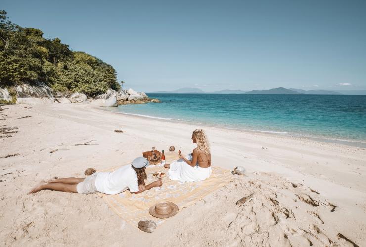 Two people picnicking on a white sand beach with the calm blue ocean in front of them on Bedarra Island, Queensland © Bedarra Island/Elise Cook