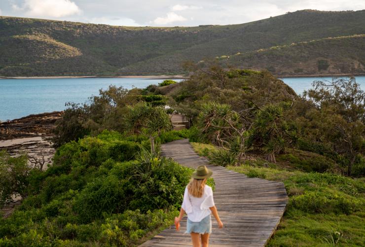 A woman walking along a wooden boardwalk surrounded by green bushland with the ocean in the distance on Pumpkin Island, Southern Great Barrier Reef, Queensland © Tourism and Events Queensland