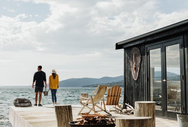 A couple standing on the edge of a balcony over the ocean beside a cottage with deck chairs and a fire pit on Satellite Island, Tasmania © Adam Gibson