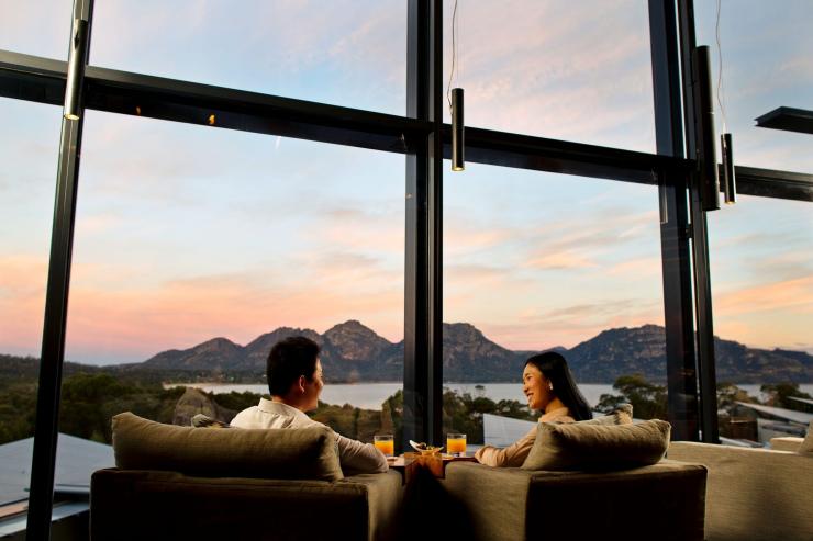A couple dining by large floor to ceiling windows overlooking a calm bay fringed by rocky mountains, Saffire Freycinet, Freycinet, Tasmania © Tourism Australia