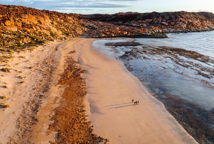 Aerial view over a group of people walking along a golden beach surrounded by towering piles of red rocks, Murujuga Aboriginal Corporation, Pilbara, Western Australia © Tourism Western Australia