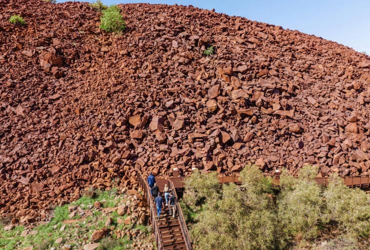 A group of people standing at the top of stairs leading to a mountain of red rocks covered in Aboriginal art, Murujuga Aboriginal Corporation, Pilbara, Western Australia © Tourism Western Australia