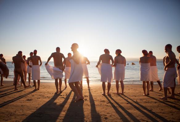A group of people wearing towels and red swim caps while standing on the beach during the early morning for the Nude Solstice Swim, Dark Mofo, Hobart Tasmania © Dark Mofo 