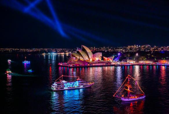 A harbour covered with rainbow boats lit up at night, and a building adorned in sails on the shore during Vivid Sydney, Sydney, New South Wales © Destination NSW