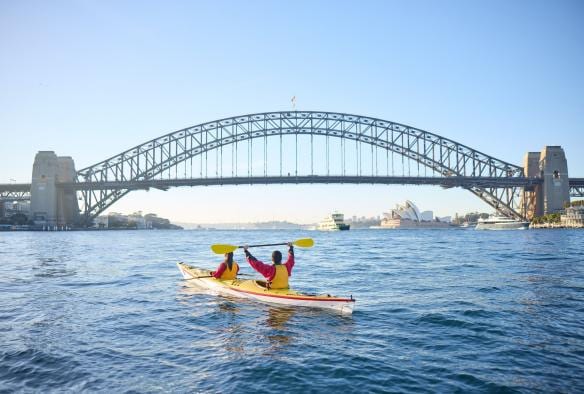 A pair of kayakers hold up their paddles in front of the Sydney Harbour Bridge in Sydney, New South Wales © Tourism Australia