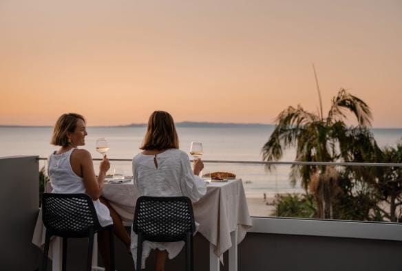 Two women face the ocean as they drink glasses of wine at Noosa Heads, Sunshine Coast, Queensland © Tourism Australia