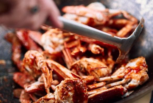 A close up photo shows a tumble of crabs being wok fried with chilli in a large, industrial sized wok. The chef’s hand is seen stirring the crab with a metal spatula. The crab looks fresh and succulent, Shuck & Shell Seafood Festival, Barrington Coast, New South Wales © Shuck & Shell Seafood Festival