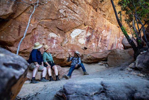 Two people sitting with a tour guide at the foot of a rocky cliff covered in rock art paintings during a tour with Jarramali Rock Art Tours, Cape York, Queensland © Tourism and Events Queensland 