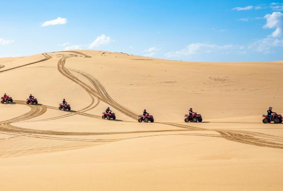 A line of people riding quad bikes across sand dunes with Sand Dune Adventures at Stockton Beach, Port Stephens, New South Wales © Destination NSW
