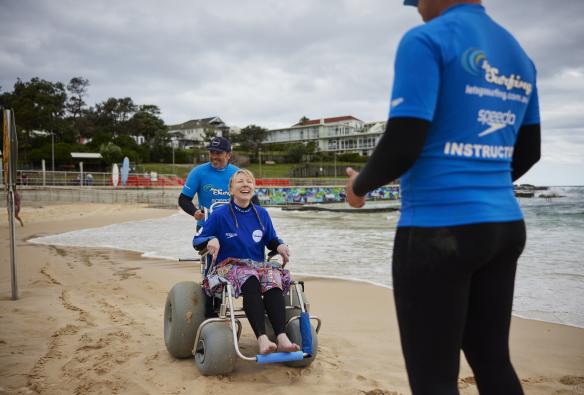 Woman in a beach wheelchair on the sand at Bondi Beach with Let's Go Surfing instructors, Sydney, NSW © Tourism Australia