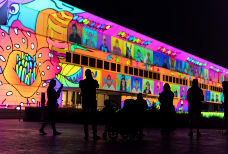 A group of people casting dark silhouettes in front of a building lit up in a colourful art installation called Illuminations, Enlighten Festival, Canberra, Australian Capital Territory © Enlighten Festival