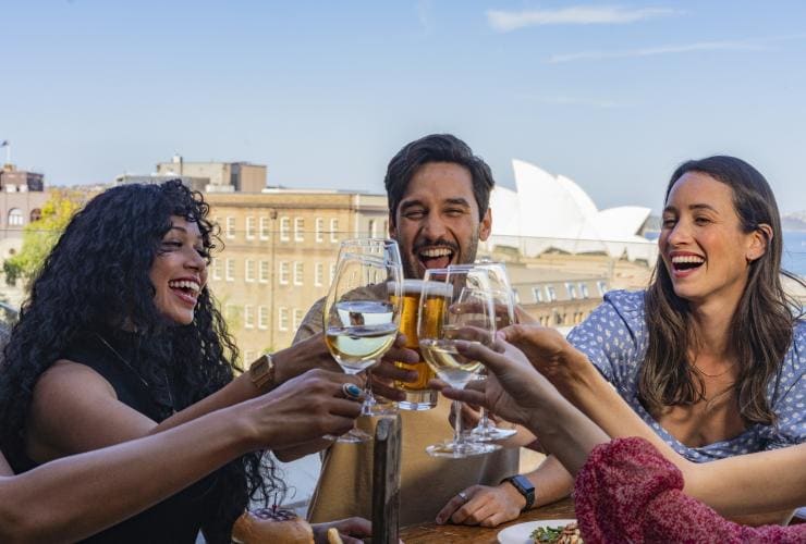 A group of friends clinking glasses on a rooftop with views of the Sydney Opera House sails behind them at The Glenmore, The Rocks, Sydney, New South Wales © Destination NSW