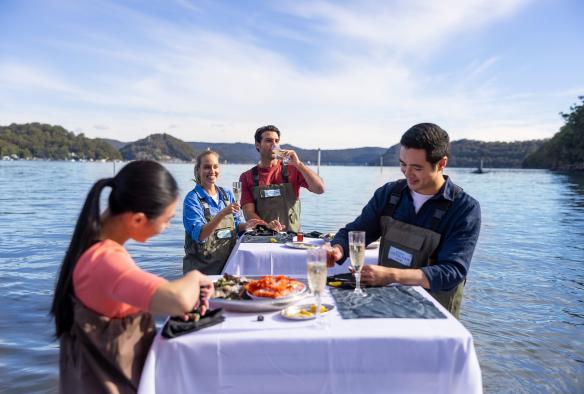 Two couples standing at tables in a river with oysters, prawns and sparkling wine at Sydney Oyster Farm Tours, Mooney Mooney, New South Wales © Tourism Australia 