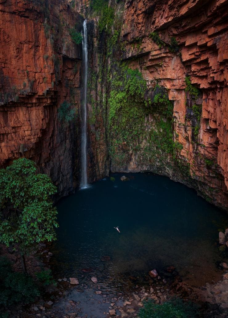 Aerial view of a person floating on their back in a deep blue waterhole as a gentle waterfall cascades down from red rock cliffs at Emma Gorge, El Questro Wilderness Park, Western Australia © Tourism Australia
