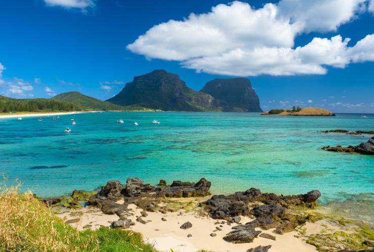Overlooking the clear blue water of Lord Howe Island dotted with boats with the island’s green mountains rising in the distance, New South Wales © Tourism Australia