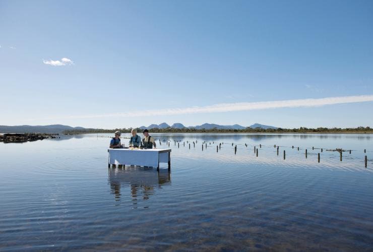 A couple dining at a table in the water at the Freycinet Marine Oyster Farm during a Saffire Signature Experience in Coles Bay, Freycinet, Tasmania © Tourism Tasmania / George Apostolidis