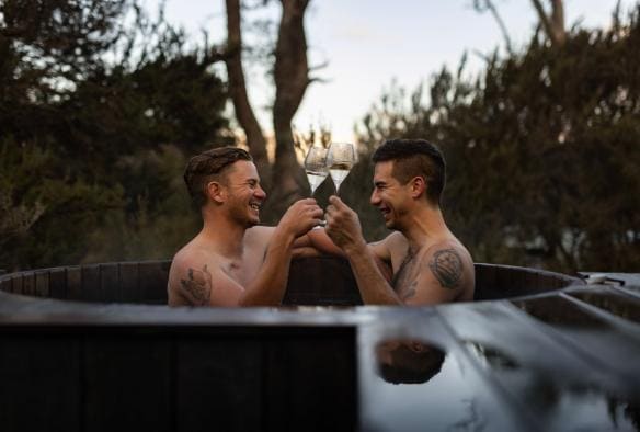 Two men smiling and drinking white wine from a wooden hot tub among the trees at Peppers Cradle Mountain Lodge in Cradle Mountain, Tasmania © Dearna Bond
