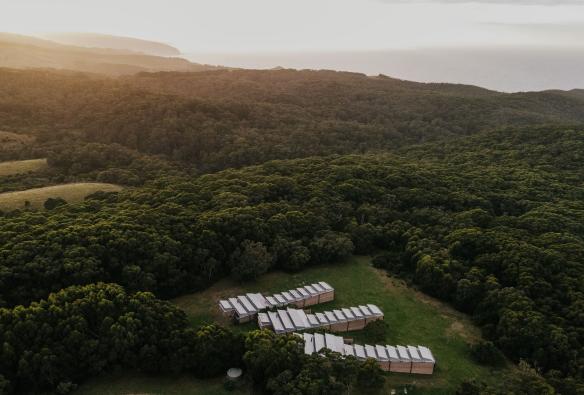 Aerial view over three rows of small buildings surrounded by greenery with the ocean visible in the distance at Alkina Lodge, Wattle Hill, Victoria © Alkina Lodge