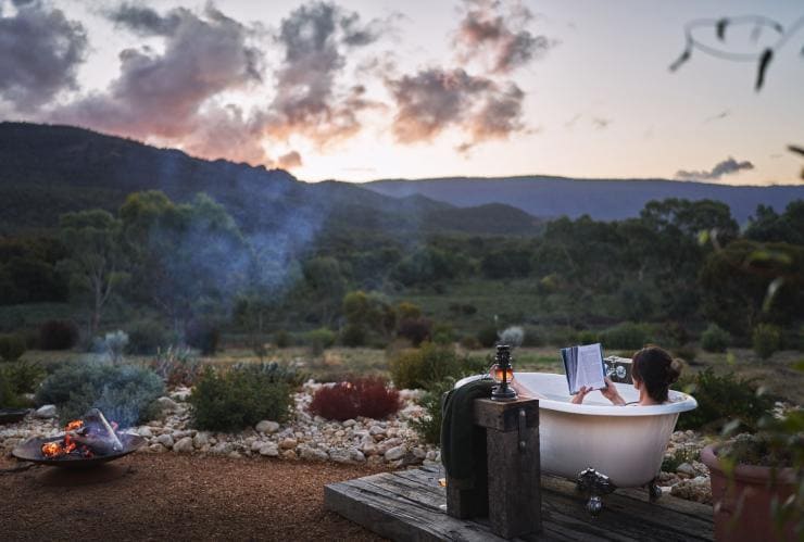 A person in an outdoor tub beside a fire pit overlooking bushland, Nook on the Hill, Grampians, Victoria © Visit Victoria