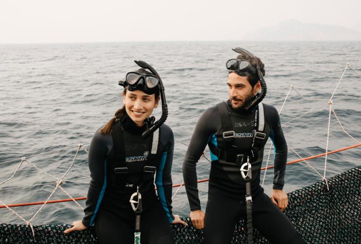 Two people sitting on the edge of a boat wearing wet suits and snorkels, Dolphin Swim Australia, Port Stephens, New South Wales © Destination NSW