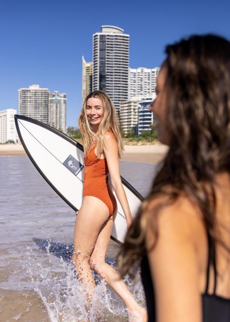 Two people walk down the sandy stretch of Surfers Paradise Beach, heading towards the ocean with surfboards in hand. The bright sun casts a golden glow over the scene, while the waves crash gently along the shore.