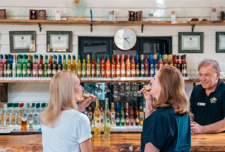 Two women sipping liqueurs at the bar in front of the bartender and a colourful display of spirits during a tour with the Vino Bus at Tamborine Mountain Distillery, Brisbane, Queensland © Tourism and Events Queensland
