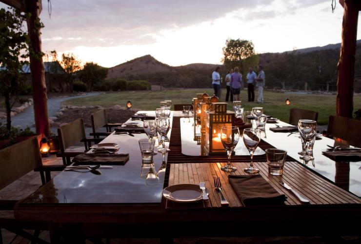 Outdoor dining table setting at dusk with a group of people standing on the grass in the background at Arkaba Conservancy, Flinders Ranges, South Australia © Arkaba Conservancy/Randy Larcombe
